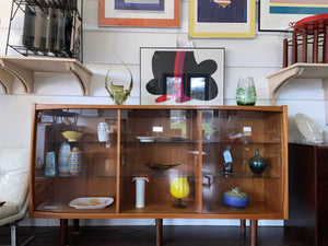 Whether to showcase your midcentury barware, vintage glassware or ceramic collection, your china or books, this gorgeous Danish designed glass front cabinet has perfect lighting to create a stunning display. Three sliding glass doors with teak trim open to adjustable height glass shelves. Subtle arch top curtain. Made in Denmark by Laurits M Larsen Mobelfabrik with interior lights off.- Cook Street Vintage