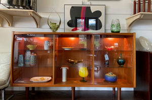 Whether to showcase your midcentury barware, vintage glassware or ceramic collection, your china or books, this gorgeous Danish designed glass front cabinet has perfect lighting to create a stunning display. Three sliding glass doors with teak trim open to adjustable height glass shelves. Subtle arch top curtain. Made in Denmark by Laurits M Larsen Mobelfabrik.- Cook Street Vintage