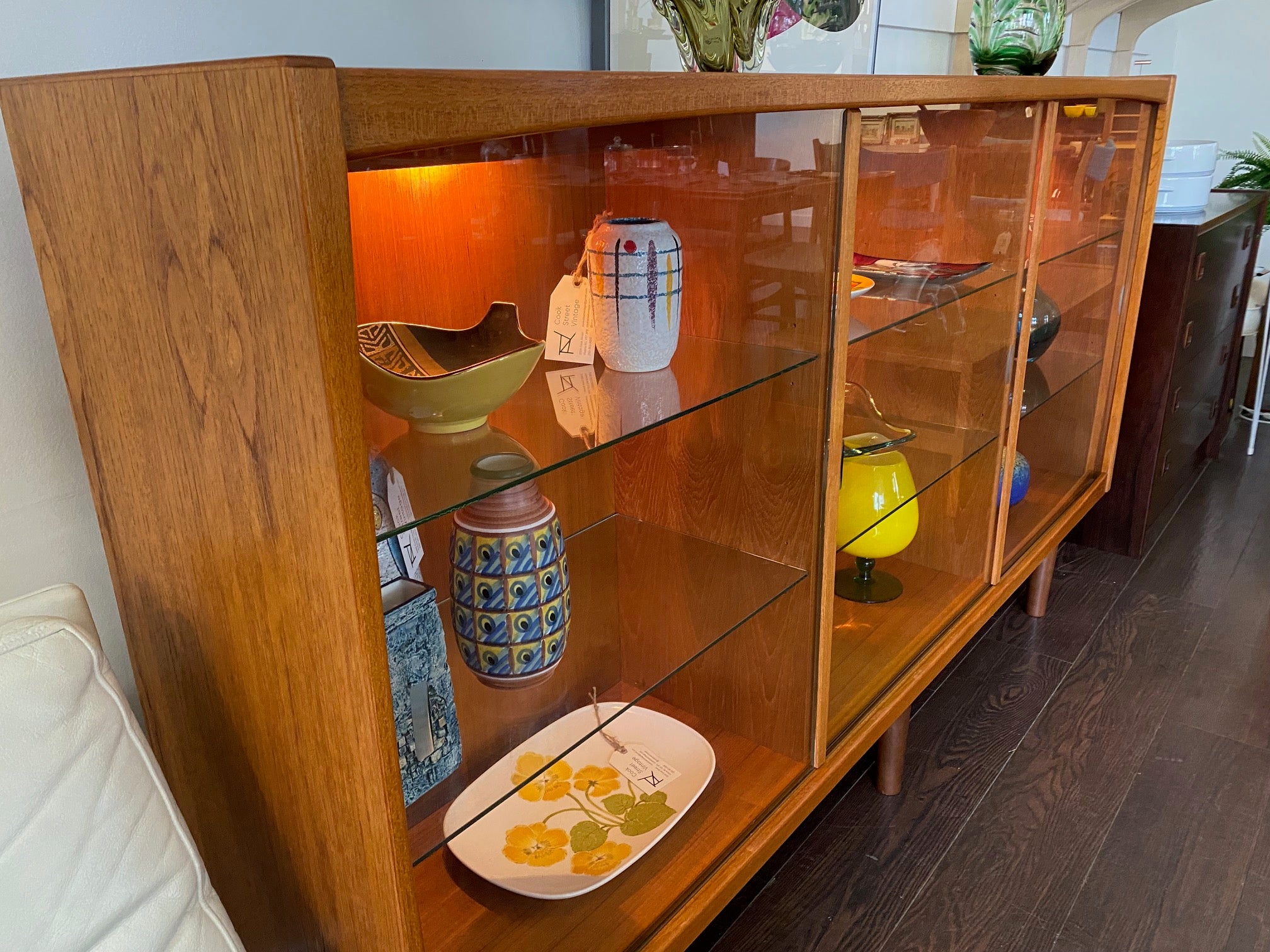 Whether to showcase your midcentury barware, vintage glassware or ceramic collection, your china or books, this gorgeous Danish designed glass front cabinet has perfect lighting to create a stunning display. Three sliding glass doors with teak trim open to adjustable height glass shelves. Subtle arch top curtain. Made in Denmark by Laurits M Larsen Mobelfabrik.- Cook Street Vintage